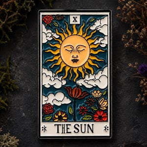 Könnte beinhalten: Eine rechteckige Emaille-Anstecknadel mit einem Tarotkarten-Motiv, das mit THE SUN und der römischen Ziffer X beschriftet ist. Die Abbildung zeigt eine gelbe Sonne mit Gesicht, weiße Wolken und bunte Blumen auf dunkelblauem Grund.