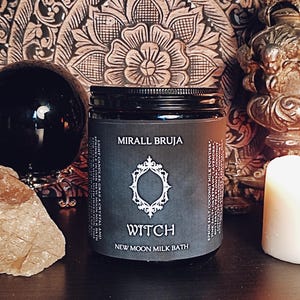 Peut inclure: Un pot noir avec l'inscription "MIRALL BRUJA WITCH NEW MOON MILK BATH" et un cadre décoratif. Le pot est posé sur une surface sombre, avec une sphère noire, un cristal et une bougie en arrière-plan. Le pot contient des ingrédients comme du sel d'Epsom, de la poudre de lait de coco et de la lavande.