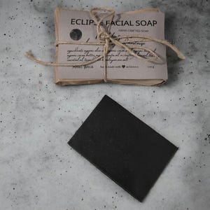 Può includere: Un sapone nero a carbone vegetale avvolto in carta marrone con spago e un'etichetta che recita "ECLIP FACIAL SOAP HAND-CRAFTED SOAP" con gli ingredienti elencati e "fatto con  in Grecia 110g"
