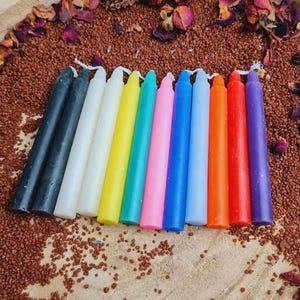 Könnte beinhalten: Ein Set mit 11 dünnen, spitz zulaufenden Kerzen in verschiedenen Farben, darunter Schwarz, Weiß, Gelb, Türkis, Rosa, Blau, Hellblau, Orange, Rot und Lila. Die Kerzen sind in einer Reihe auf einer braunen Oberfläche angeordnet, auf der rote und braune samenartige Objekte verstreut sind.