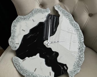 May include: A decorative wall clock with a unique design. The clock face is split into sections of white, black, and marble-like patterns. The edges are adorned with silver glitter, and the clock hands are silver.