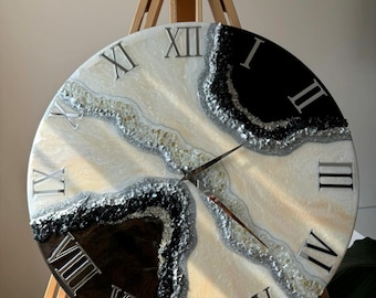 May include: Large, round wall clock with a cream, black, and silver design. The clock face features Roman numerals and a unique, textured pattern. The clock is displayed on a wooden easel.