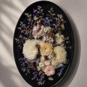May include: An oval black tray featuring an arrangement of dried flowers in shades of cream, pale pink, and deep purple. The composition includes large, textured blooms, delicate sprigs of baby's breath, and small purple filler flowers.