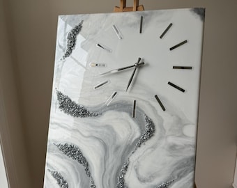 May include: A rectangular wall clock with a white and gray marbled design. The clock features silver-colored hour markers and hands. Decorative silver-colored glitter accents the marbled pattern. The clock is displayed on a wooden easel.