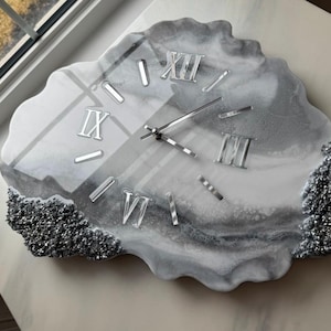 Può includere: Un orologio da parete dalla forma irregolare ispirata a una geode, con una finitura lucida nei toni del bianco, grigio e nero. Il quadrante presenta numeri romani argentati e lancette sottili, arricchito da dettagli in glitter argento.