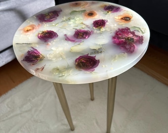 May include: A white round table with a clear epoxy top featuring pressed flowers and leaves. The table has three gold metal legs.