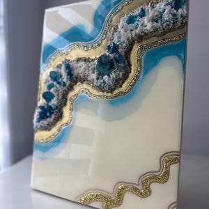Könnte beinhalten: Abstrakte Kunst aus Kunstharz mit einem Geoden-Design. Das Kunstwerk hat eine weiße Basis mit blauen und goldenen Akzenten sowie strukturierte Bereiche mit blauen Kristallen. Die Kanten sind mit goldenem Glitzer versehen.