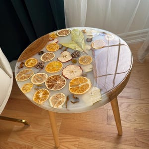 Peut inclure: Une table d'appoint ronde avec un dessus en résine transparente incrusté de tranches d'orange et de pomme séchées, et un bord en bois. La table a trois pieds en bois de couleur claire. La table mesure environ 51 cm de diamètre.