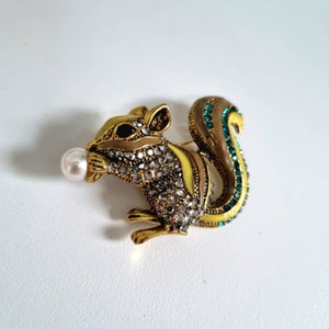 May include: A gold-tone squirrel brooch with a pearl on its mouth. The squirrel is decorated with rhinestones and green enamel accents.