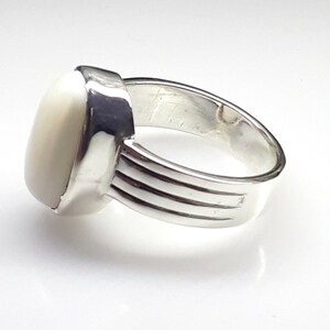 May include: A silver ring with a large, oval, off-white stone. The ring has a wide band with three horizontal grooves. The stone is set in a square bezel. The ring is shown on a white background.
