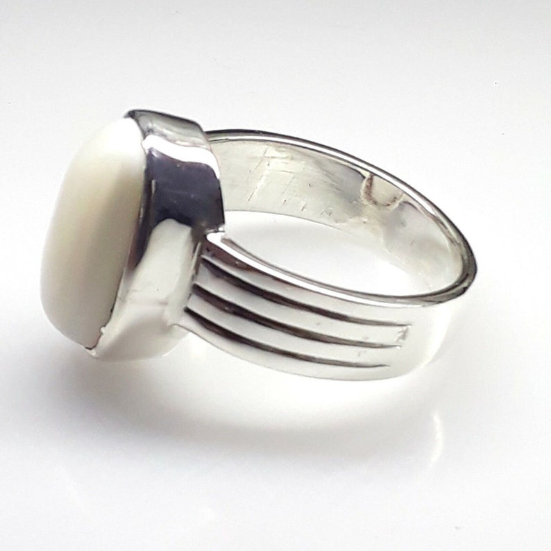 May include: A silver ring with a large, oval, off-white stone. The ring has a wide band with three horizontal grooves. The stone is set in a square bezel. The ring is shown on a white background.