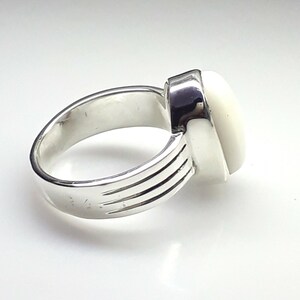 May include: A silver ring with a large, oval, off-white stone. The ring band has three parallel grooves on the side. The setting is a simple, modern design, showcasing the stone's shape and color.