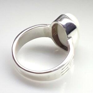 May include: A silver ring with a large, oval, off-white stone. The ring's band is thick and polished, with subtle etched lines on the sides. The stone is set in a smooth, bezel setting, creating a classic and elegant look.