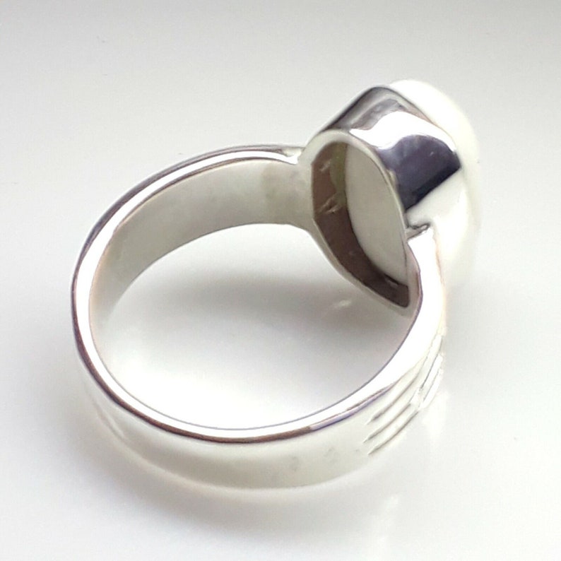 May include: A silver ring with a large, oval, off-white stone. The ring's band is thick and polished, with subtle etched lines on the sides. The stone is set in a smooth, bezel setting, creating a classic and elegant look.