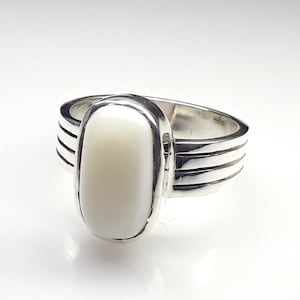May include: A silver ring featuring a large, oval, creamy white stone. The band has three parallel grooves on each side, adding a textured detail. The ring's design is simple yet elegant, suitable for everyday wear or special occasions.