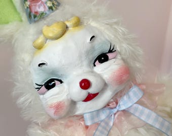 May include: A vintage-style plush bunny with a painted face, wearing a party hat with floral accents and a pink and blue gingham bow. The bunny has a red nose, rosy cheeks, and a ruffled collar. The plush is white with a fluffy texture.