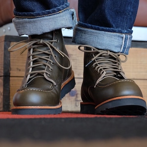 May include: A pair of olive green leather boots featuring a moccasin-style toe, brown laces, and a thick black lug sole. The boots have a smooth finish with visible stitching along the toe box and a natural tan leather midsole layer.