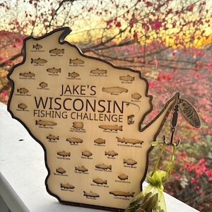 May include: Wooden Wisconsin fishing challenge sign with various fish illustrations and the text "Jake's Wisconsin Fishing Challenge." A small green bag with fish-shaped charms hangs from the sign. The background features fall foliage.