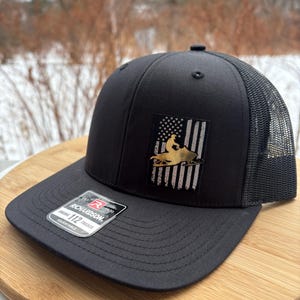 May include: Black trucker hat with a mesh back and a flat brim. The hat features a patch with an American flag design and a gold-colored snowmobile graphic. The brand tag "Richardson 112 Trucker" is visible on the front.