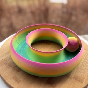 Pode incluir: Uma tigela decorativa em espiral multicolorida com uma bola. Apresenta um gradiente de cores rosa, amarelo, verde e azul. A tigela está colocada sobre uma superfície de madeira, criando um design vibrante e atraente.