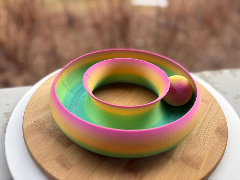Peut inclure: Un bol d&eacute;coratif en spirale multicolore avec une boule. Il pr&eacute;sente un d&eacute;grad&eacute; de rose, jaune, vert et bleu. Le bol est pos&eacute; sur une surface en bois, cr&eacute;ant un design vibrant et attrayant.