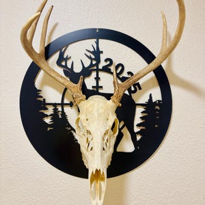 May include: A deer skull with large antlers is mounted on a black metal wall art piece. The art features a silhouette of a deer, trees, and the numbers "2023" within a circular design. The skull is off-white.