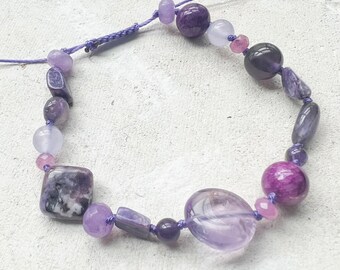 May include: A handmade purple gemstone bracelet featuring a variety of beads in shades of lavender, amethyst, and deep violet. The beads include round, oval, and square shapes, strung on a purple cord. The bracelet is designed for adjustable sizing.