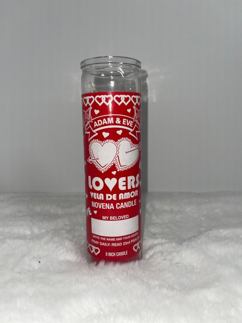 Może przedstawiać: Przezroczysta szklana świeca z czerwoną etykietą z napisem "ADAM & EVE" i "LOVERS VELA DE AMOR NOVENA CANDLE". Etykieta zawiera motywy serca i miejsce na wpisanie imienia. Świeca ma około 20 cm wysokości.