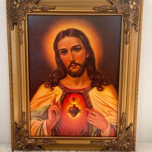 May include: A framed painting of Jesus Christ with a golden frame. The painting depicts Jesus with his hands over his heart, which is on fire and surrounded by thorns. The background is a dark red with a golden halo around Jesus' head.