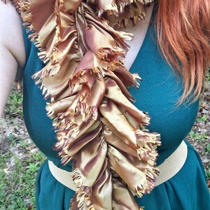 May include: A long, ruffled scarf in a shimmering bronze and copper tone, featuring frayed, raw-edged fabric trim along the entire length. The scarf is draped over a dark teal, textured sleeveless dress with a wide, light beige elastic waistband.