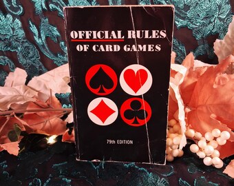 May include: A black paperback book titled Official Rules of Card Games 79th Edition. The cover features white text and four circular icons representing card suits: a black spade, a red heart, a red diamond, and a black club on a black background.