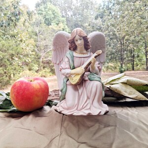 May include: A ceramic angel figurine with light brown hair and large wings, wearing a pale pink robe with green accents, playing a small stringed lute. Beside the angel sits a large red and yellow apple and a cluster of dried brown leaves.