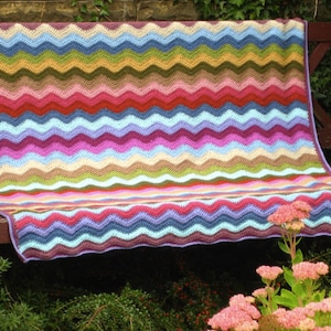 May include: A colorful crocheted blanket with a chevron pattern in shades of pink, blue, green, and brown. The blanket is draped over a wooden bench.