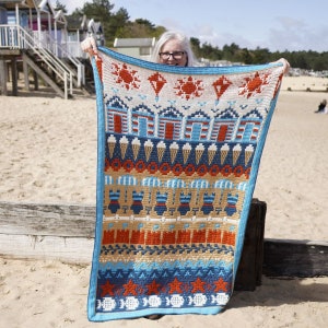 May include: A colourful crochet blanket with a beach theme. The blanket features a variety of designs, including beach houses, ice cream cones, and seashells. The blanket is made with a variety of colours, including blue, red, white, and yellow.