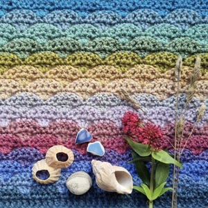 May include: A crocheted blanket in a gradient of blues, greens, yellows, and pinks, resembling ocean waves. The blanket is adorned with seashells, broken pottery, a flower, and dried grass. The handmade item evokes a coastal theme.
