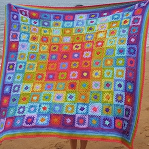 May include: A large, colorful, crocheted blanket with a square pattern. The blanket features a vibrant rainbow of colors, including red, orange, yellow, green, blue, and purple. The blanket is held up against a beach background.