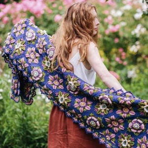 Puede incluir: Un chal azul tejido a crochet con patrones florales en rosa, verde y blanco. El chal está drapeado sobre los hombros, mostrando el intrincado diseño hecho a mano. El fondo presenta un jardín con flores rosas y follaje verde.