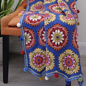 May include: A vibrant, handmade crochet blanket with a blue background and colorful floral patterns in red, yellow, and white. The blanket is adorned with pom-poms in various colors along the edges. The blanket is draped over an orange chair.