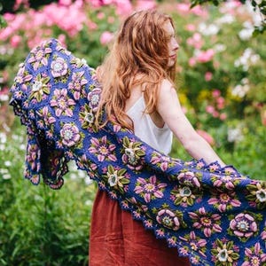 May include: A blue crochet shawl with pink, green, and cream floral patterns. The shawl is displayed, highlighting the detailed crochet work. The background shows a garden with pink flowers, creating a vibrant contrast.