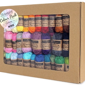 Puede incluir: Una caja de 109 ovillos de hilo Catona Scheepjes en un arcoíris de colores. La caja es de cartón marrón y tiene una ventana de plástico transparente.