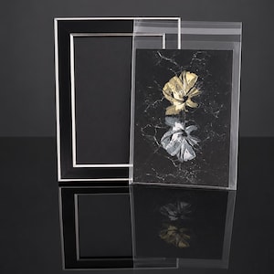 May include: A rectangular black picture frame with a thin silver inner border, positioned next to a clear plastic sleeve containing a black card with a marble texture and two stylized flower illustrations in gold and silver foil.