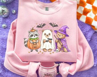 May include: A light pink crewneck sweatshirt featuring a graphic of a raccoon in a pumpkin costume, a bear dressed as a ghost, and a bear wearing a purple witch hat and cape holding a broom, with small bats and candy corn scattered around.