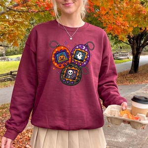 May include: A maroon crewneck sweatshirt featuring a graphic print of three digital pet keychains. The orange, purple, and black keychains display pixelated images of a jack-o'-lantern, a white ghost, and a skeleton making a peace sign.