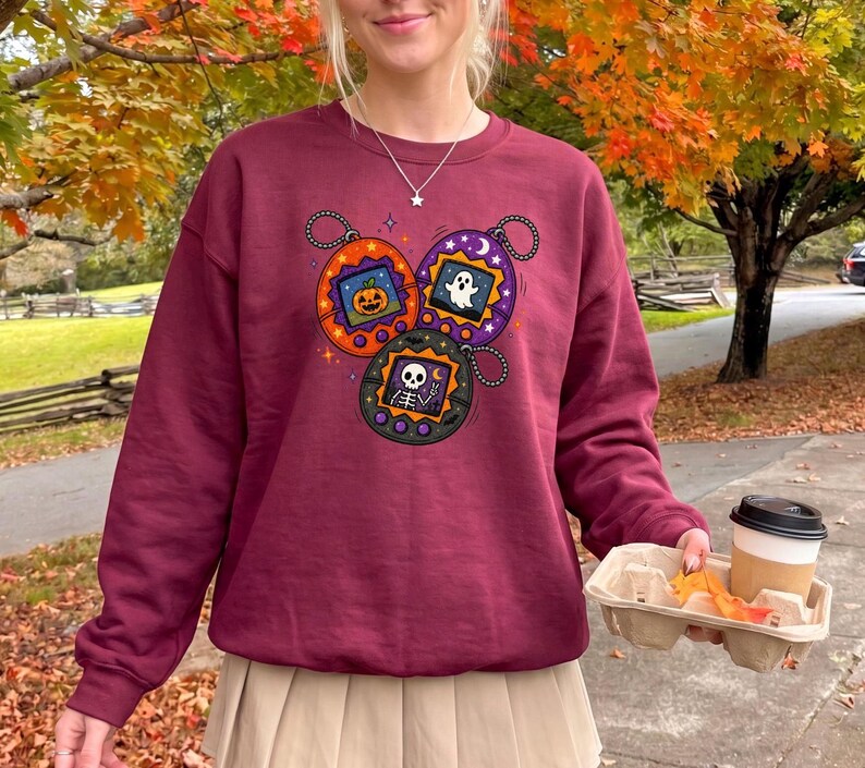 May include: A maroon crewneck sweatshirt featuring a graphic print of three digital pet keychains. The orange, purple, and black keychains display pixelated images of a jack-o'-lantern, a white ghost, and a skeleton making a peace sign.
