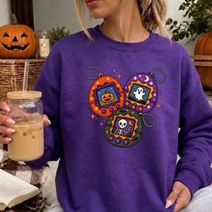 May include: A purple long-sleeve sweatshirt featuring a graphic print of three digital pet keychains. The orange, purple, and black keychains display pixelated images of a jack-o'-lantern, a ghost, and a skeleton making a peace sign.