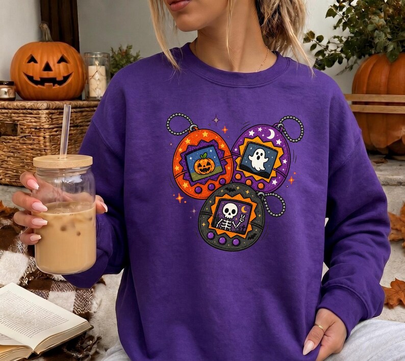 May include: A purple long-sleeve sweatshirt featuring a graphic print of three digital pet keychains. The orange, purple, and black keychains display pixelated images of a jack-o'-lantern, a ghost, and a skeleton making a peace sign.