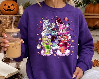 May include: A purple long-sleeve sweatshirt featuring a graphic print of five colorful cartoon bears dressed in Halloween costumes, including a mummy, a vampire, and a Frankenstein monster, surrounded by small stars, bats, and pumpkins.