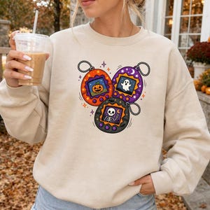 May include: A beige crewneck sweatshirt featuring a graphic print of three round digital pet toys. The orange toy displays a jack-o'-lantern, the purple toy shows a ghost, and the black toy depicts a skeleton making a peace sign with its hand.