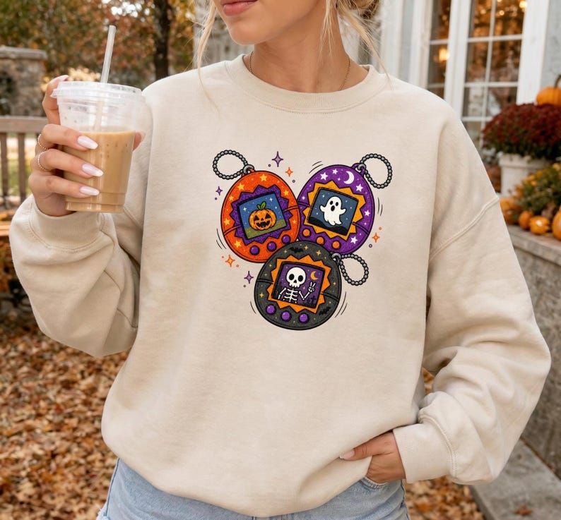 May include: A beige crewneck sweatshirt featuring a graphic print of three round digital pet toys. The orange toy displays a jack-o'-lantern, the purple toy shows a ghost, and the black toy depicts a skeleton making a peace sign with its hand.