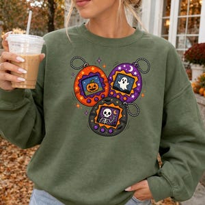 May include: An olive green crewneck sweatshirt featuring a graphic print of three digital pet keychains. The orange, purple, and black devices display a jack-o'-lantern, a white ghost, and a skeleton respectively, accented by small stars and moons.